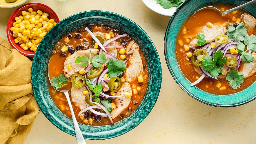 Zwei Schalen mit einer herzhaften Suppe, die Stücke von Hühnerfleisch, schwarze Bohnen, Mais, rote Zwiebeln und Jalapeños enthält. Frische Korianderblätter garnieren die Suppe, die in hellen, geflammten Schalen serviert wird. Neben der Suppe steht eine Schale mit Maiskernen.