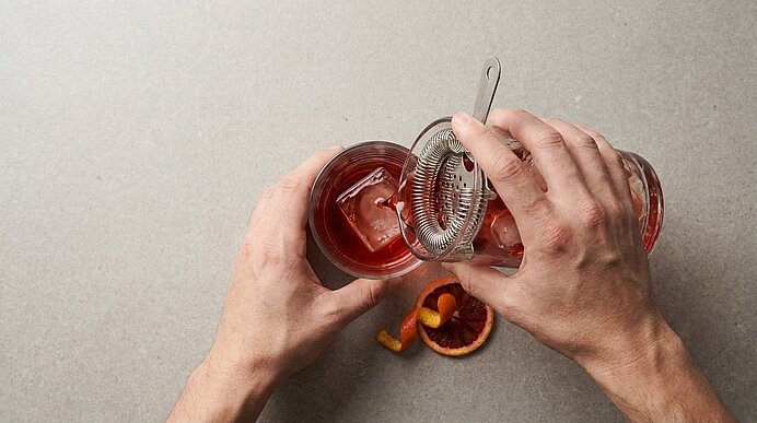 Zwei Hände gießen einen roten Cocktail durch ein Barsieb in ein mit Eis gefülltes Glas; daneben Orangenschale und eine halbierte Blutorange.