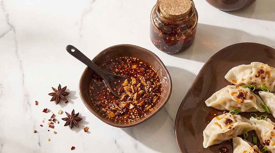 Schüssel mit scharfem Chili-Öl und Löffel neben Teller mit gedämpften Teigtaschen (Dumplings); Glas Chilipaste, Sternanis und Chiliflocken auf hellem Marmor.