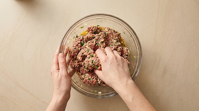Zwei Hände kneten gewürztes Hackfleisch mit Kräutern in einer Glasschüssel.