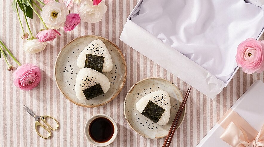 Zwei dreieckige Onigiri-Reisbällchen mit Sesam und Nori auf Keramiktellern, daneben Chopsticks und eine kleine Schale Sojasauce auf gestreiftem Tuch.