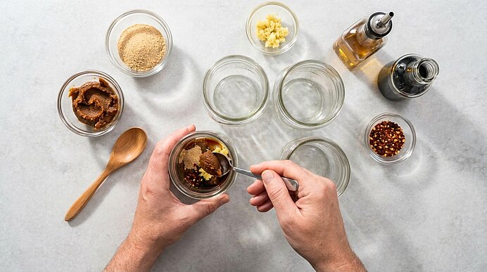 Hände rühren in einem Einmachglas eine Würzpaste, umgeben von kleinen Schalen mit braunem Zucker, gehacktem Knoblauch, Chiliflocken sowie Öl- und Essigflaschen.