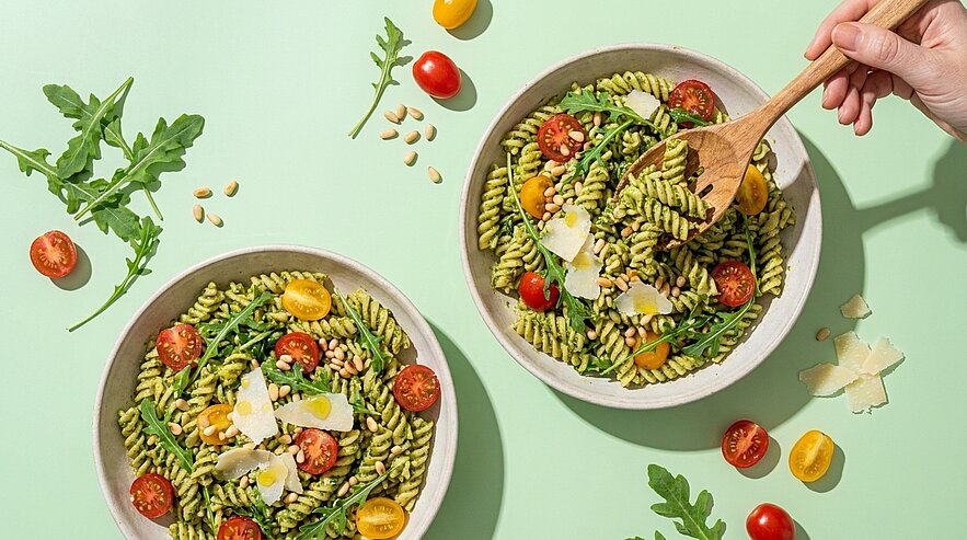 Zwei Schalen Fusilli mit grünem Pesto, Rucola, halbierten roten und gelben Kirschtomaten, Pinienkernen und Parmesanspänen; rechts eine Hand rührt mit einem Holzlöffel.