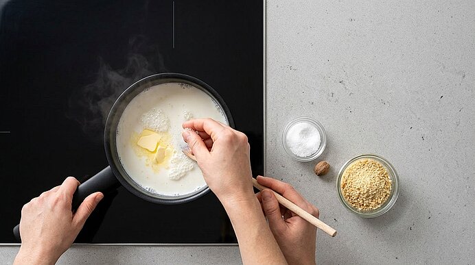Hände würzen heiße Milch mit Butter in einem Stieltopf auf dem schwarzen Kochfeld.