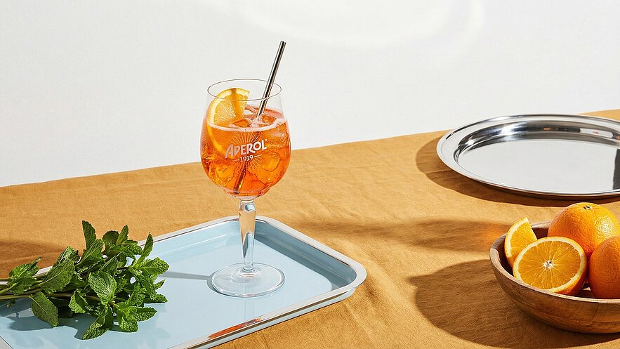 Ein Glas Aperol-Drink mit Eis und einer Orangenscheibe steht auf einem blauen Tablett. Daneben liegen frische Minzblätter. Im Hintergrund sind ein silbernes Tablett und eine Schüssel mit verschiedenen Orangenschnitten auf einem braunen Tisch sichtbar.