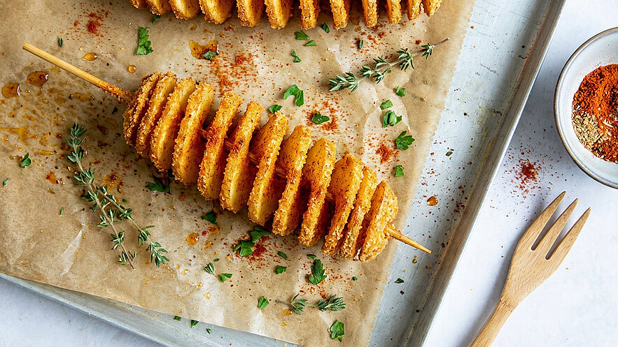Auf einem Blech liegen spiralig aufgereihte Kartoffelscheiben, die goldbraun frittiert sind. Sie sind mit Gewürzen bestreut und umgeben von frischen Kräutern wie Thymian und Petersilie. Ein kleiner Teller mit Gewürzen ist ebenfalls sichtbar.