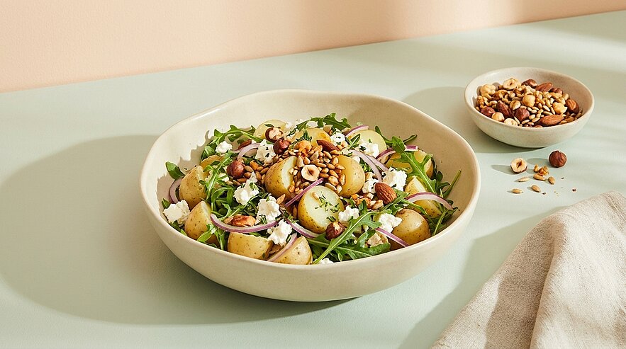 Schüssel mit Kartoffelsalat aus halbierten kleinen Kartoffeln, Rucola, roten Zwiebelringen, zerbröseltem Käse und gerösteten Haselnüssen; daneben eine kleine Schale mit Nüssen.