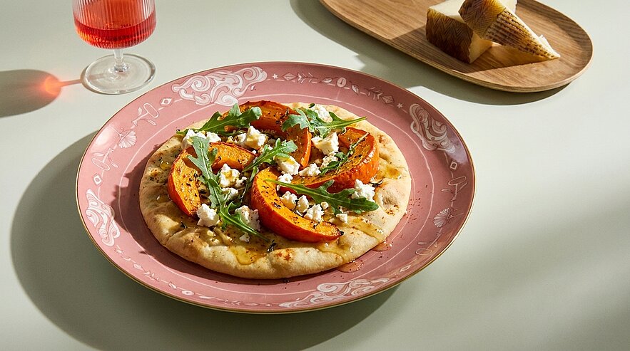 Fladenbrot mit gegrillten Pfirsichscheiben, Rucola und zerbröseltem Feta, auf einem dekorativen rosa Teller angerichtet.