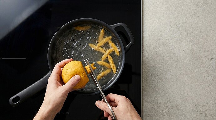 Hände schneiden mit einer Schere Zitronenschale in Streifen über einem kochenden Topf; die Streifen fallen ins Wasser.