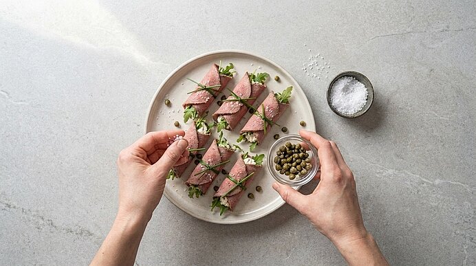 Zwei Hände bestreuen angerichtete Roastbeef-Röllchen auf einer Platte mit Flockensalz und Kapern.