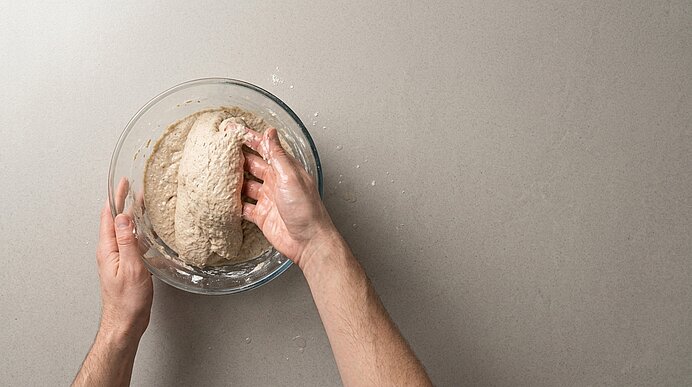 Zwei Hände formen den klebrigen Roggenteig in einer Glasschüssel auf einer hellgrauen Steinarbeitsplatte.