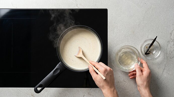 Zwei Hände rühren mit einem Holzlöffel eine cremige, helle Sauce in einem Topf auf dem Herd; rechts zwei kleine Schälchen mit Flüssigkeit und einer Vanilleschote.
