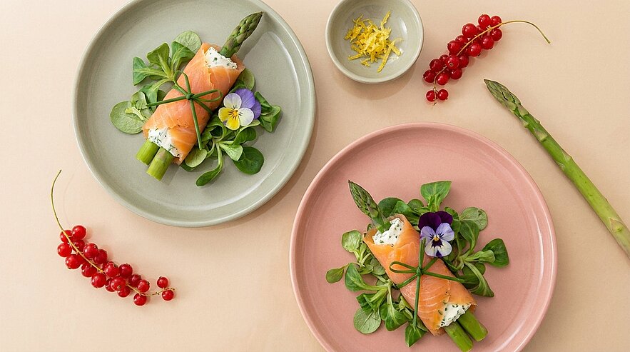 Zwei Teller mit je drei grünen Spargelstangen, in Räucherlachsscheiben gewickelt und mit Schnittlauchbändern, auf Feldsalat und mit essbaren Blüten garniert.