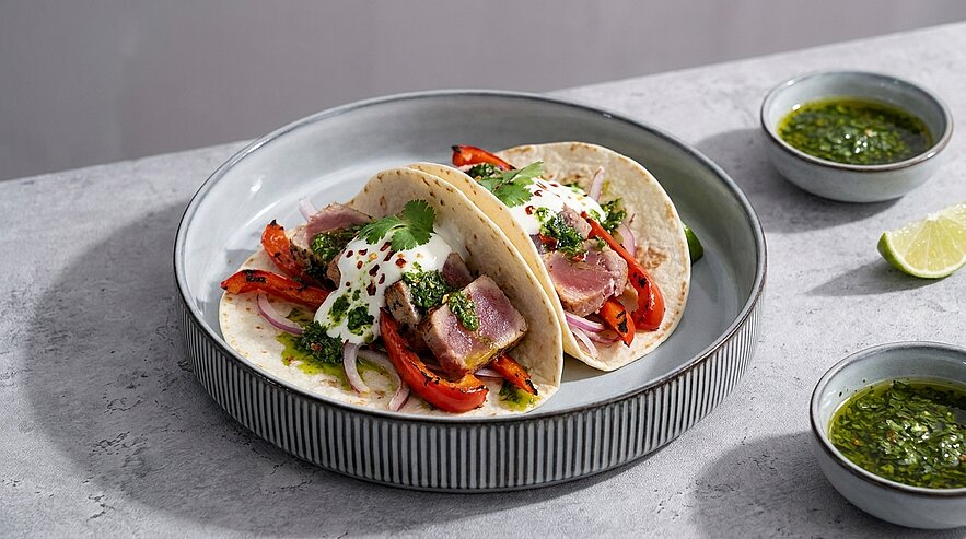 Zwei weiche Tacos mit kurz angebratenem Thunfisch, gegrillter roter Paprika, Zwiebeln, frischem Koriander und cremiger Sauce; dazu zwei Schälchen grüner Kräutersauce und eine Limettenspalte.