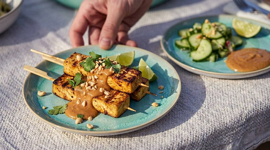Auf einem Teller liegen gegrillte, gewürzte Tofu-Spieße, garniert mit Erdnusssauce, Koriander und Limettenscheiben. Im Hintergrund befindet sich eine Schüssel mit einem frischen Gurkensalat. Ein Hand greift nach den Spießen.