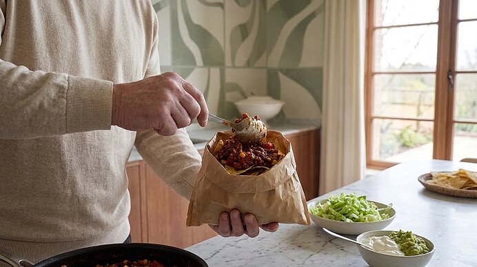 Hände einer Person schöpfen Chili mit Bohnen aus einer Pfanne in eine braune Papiertüte; auf der Arbeitsplatte stehen Schüsseln mit gehacktem Salat, Sauerrahm und Guacamole sowie Tortillachips.