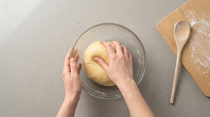 Zwei Hände formen einen runden Hefeteig in einer Glasschüssel auf einer Arbeitsfläche; daneben Schneidebrett und Holzlöffel mit etwas Mehl.