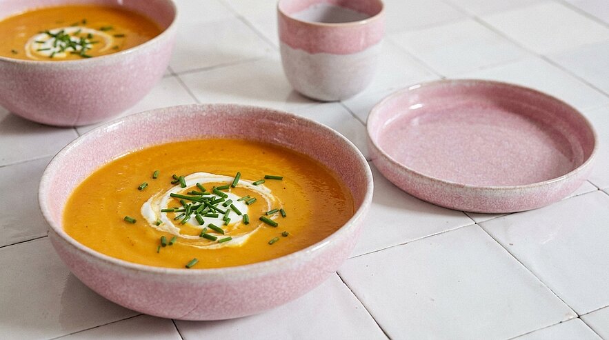 Cremige orangefarbene Suppe in einer pastellrosa Schüssel, garniert mit einem Sahneklecks und gehacktem Schnittlauch; im Hintergrund weitere rosa Keramikschalen und eine zweite Suppe.