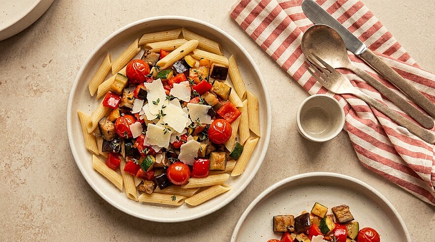 Schüssel Penne mit gebratenen Auberginenwürfeln, Kirschtomaten, frischem Basilikum und Parmesanspänen.