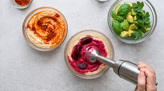 Nahaufnahme von drei Dips: links Paprika-Hummus, Mitte Rote-Bete-Hummus, der mit einem Stabmixer püriert wird, rechts Avocado mit Basilikum und Petersilie.