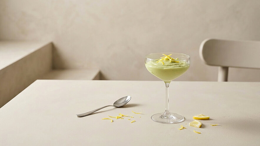 Ein elegantes Glass mit einer cremigen, grünen Nachspeise, garniert mit Zitronenzesten. Neben dem Glass liegt ein silberner Löffel. Auf dem Tisch sind einige Zitronenstücke und Zesten verstreut. Der Hintergrund ist neutral und minimalistisch gestaltet.