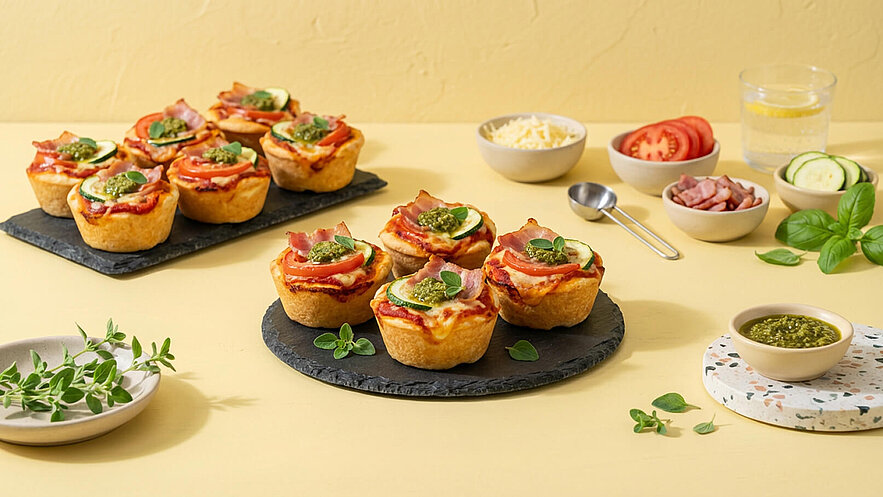 Auf einem schwarzen Platten liegen mehrere Mini-Pizza-Formen mit Tomatensauce, Käse, Zucchini, Schinken und Pesto. Im Hintergrund befinden sich kleinere Schalen mit Zutaten wie Käse, Tomaten und Gurken sowie ein Glas Wasser mit Zitrone. Der Hintergrund ist hellgelb.