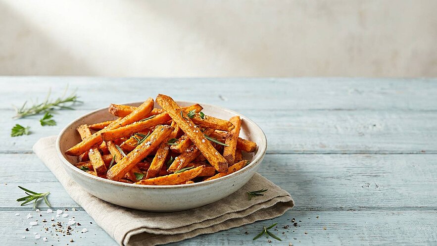 Eine weiße Schüssel mit knusprigen Süßkartoffelpommes steht auf einem hellblauen Tisch. Die Pommes sind goldbraun und mit Kräutern bestreut. Im Hintergrund sind einige grüne Kräuterblätter leicht sichtbar.