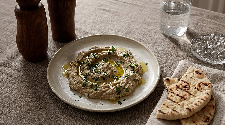 Ein Teller mit cremigem Hummus, garniert mit frischen Kräutern und einem Spritzer Olivenöl. Neben dem Hummus liegt ein Stück gegrilltes Fladenbrot. Der Hintergrund zeigt eine elegant gedeckte Tischoberfläche.