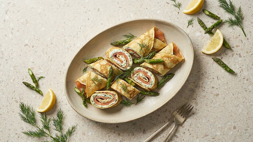 Auf einem ovalen Teller sind geschnittene Pfannkuchen-Rollen gefüllt mit Lachs und Kräutercreme angerichtet. Neben den Rollen liegen frische grüne Spargelstücke und einige Zitronenscheiben, die dem Gericht eine frische Note verleihen.