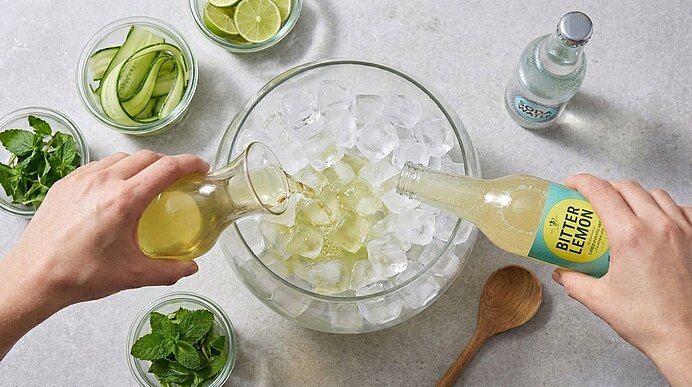 Zwei Hände gießen Holunderansatz und Bitter Lemon in eine Glasschüssel voller Eiswürfel auf heller Arbeitsplatte.