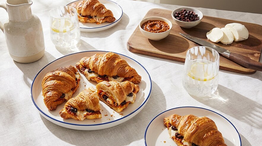 Drei Teller mit frischen, goldbraunen Croissants, einige aufgeschnitten und mit Marmelade und Käse gefüllt, angerichtet auf hellem Tisch.