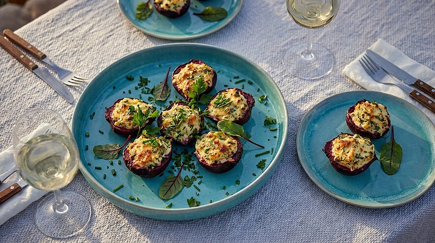Eine große blaue Schale mit gebackenen Mini-Quiches, garniert mit frischem Schnittlauch und Spinatblättern. Daneben kleinere Teller mit ähnlichen Quiches. Im Hintergrund sind Gläser mit leichtem Wein zu sehen, auf einer gedeckten Tischdecke.