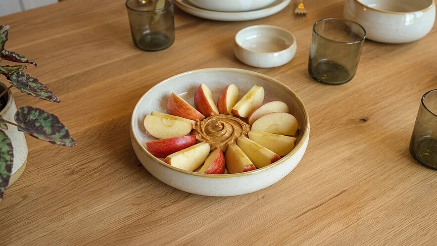 Eine flache Schale mit aufgeschnittenen Äpfeln in roten und grünen Farben. In der Mitte befindet sich ein cremiger Dip. Die Schale steht auf einem Holztablett, umgeben von kleinen Gläsern.
