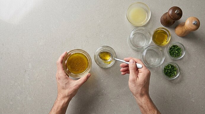 Zwei Hände rühren mit einem Löffel eine Vinaigrette in einer kleinen Glasschale; daneben Schälchen mit Olivenöl, Zitronensaft und gehackten Kräutern sowie Salz- und Pfeffermühlen.
