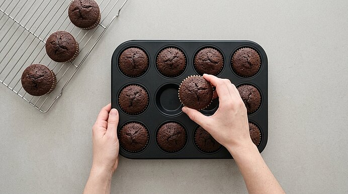 Zwei Hände heben gebackene Schokoladen-Muffins aus einer schwarzen Muffinform auf ein Kuchengitter.