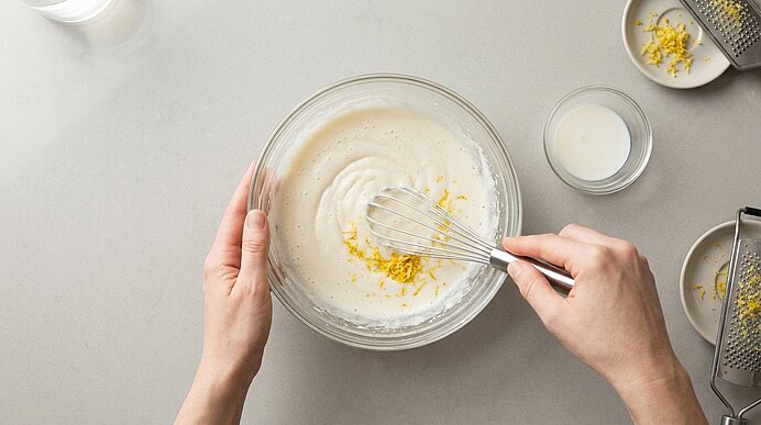 Hände schlagen mit einem Schneebesen eine helle Creme in einer Glasschüssel, bestreut mit geriebener Zitronenschale.