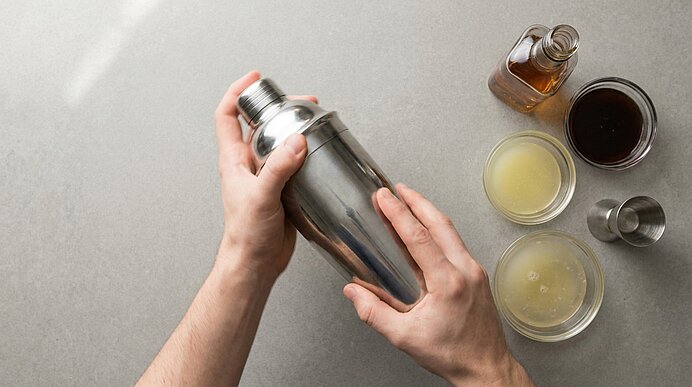 Zwei Hände schütteln einen Metall-Cocktailshaker; daneben kleine Schälchen mit Zitronensaft, Sirup, Likör und ein Messbecher.