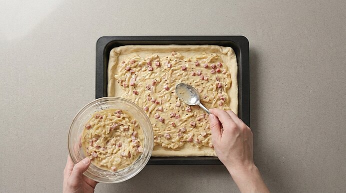 Zwei Hände verteilen die Zwiebel-Schmand-Masse mit einem Löffel aus einer Glasschüssel auf dem Teig in der Backform.