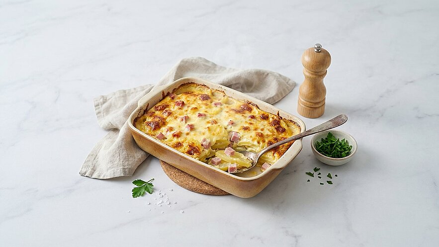 Ein frisch gebackenes Gericht aus Kartoffeln, Schinken und Käse in einer Auflaufform. Es ist golden und knusprig oben, und ein Löffel ist in der Auflaufform platziert. Neben der Form liegt ein kleiner Teller mit frisch gehacktem Schnittlauch.