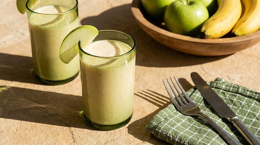Zwei Gläser grüner Smoothie stehen auf einem Tisch, verziert mit einem Apfelstück. Im Hintergrund befindet sich eine Schüssel mit grünen Äpfeln und Bananen. Daneben liegt ein kariertes Geschirrtuch mit Besteck.