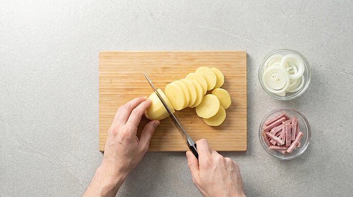 Zwei Hände schneiden eine Kartoffel mit Messer in dünne Scheiben auf einem Holzbrett; daneben zwei Schälchen mit Zwiebelringen und Schinkenstreifen.