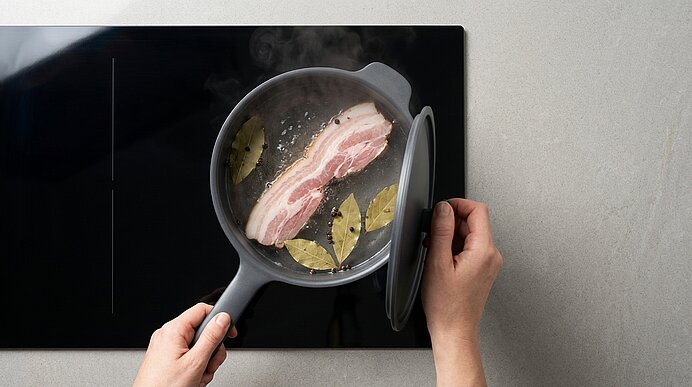 Hände halten Pfanne und Deckel; ein dicker Speckstreifen brät in der Pfanne mit Lorbeerblättern, Dampf steigt.