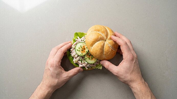 Zwei Hände legen die obere Hälfte auf das mit Fleischsalat, Gurkenscheiben und Schnittlauch belegte Brötchen.