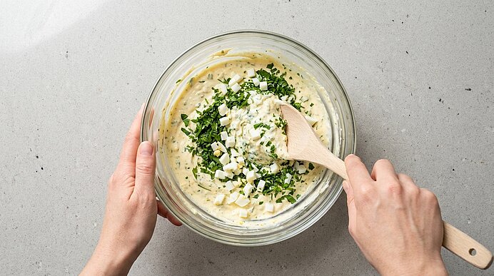 Zwei Hände rühren in einer Glasschüssel cremigen Kräuterquark/Dip mit fein gehackten Kräutern, gerührt mit einem Holzlöffel.