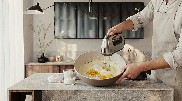 Person rührt mit elektrischem Handmixer Eier und Teig in einer großen Rührschüssel auf einer Küchentheke.