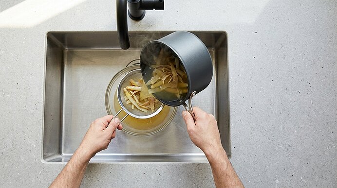 Hände gießen gekochte Pasta und heißes Kochwasser aus einem Topf durch ein Sieb in eine Glasschüssel über einem Spülbecken.