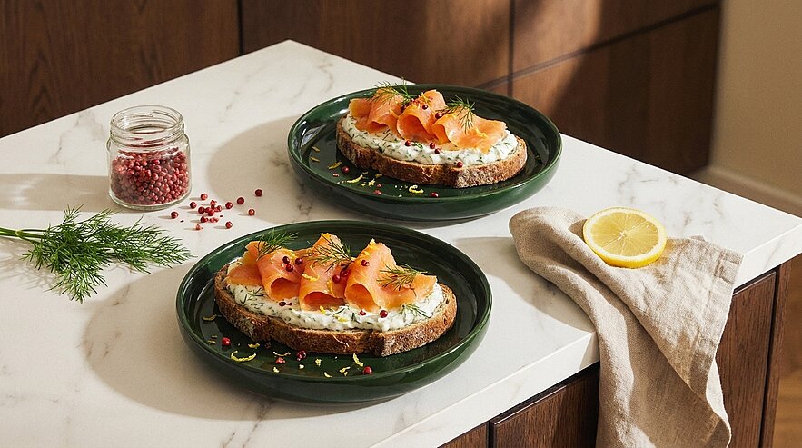 Zwei offene Brote auf dunkelgrünen Tellern mit Frischkäse, geräuchertem Lachs, Dill und rosa Pfefferkörnern; daneben eine Zitronenscheibe auf einer Serviette und ein kleines Glas Pfeffer.