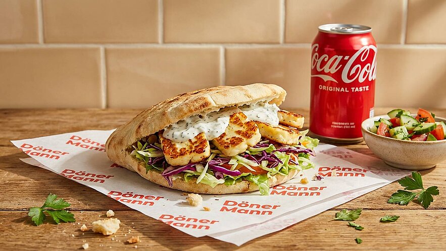 Ein frisch zubereiteter Döner im Fladenbrot, gefüllt mit Salat, Rotkohl und gebratenem Hähnchen. Neben dem Döner steht ein Glas mit Coca-Cola und eine kleine Schüssel mit gemischtem Salat. Der Hintergrund ist hell und die Präsentation ist ansprechend.