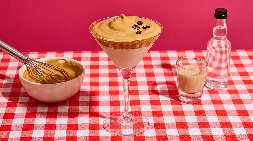 Cocktailglas mit cremigem, aufgeschlagenem Kaffeeschaum (Dalgona-ähnlich), garniert mit drei Kaffeebohnen, auf rot-weiß kariertem Tischtuch.