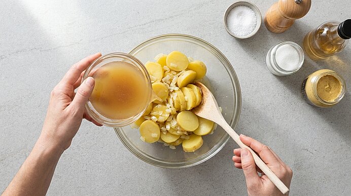 Zubereitung eines Kartoffelsalats: Hände gießen Dressing über in Scheiben geschnittene, gekochte Kartoffeln in einer Glasschüssel, ein Holzlöffel rührt, Gewürze und Öl stehen daneben.